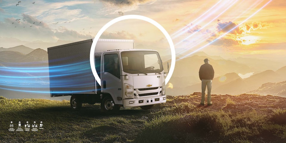 Camión Chevrolet NHR con tecnología OnStar, estacionado en un paisaje montañoso al atardecer junto a un hombre contemplando la vista.